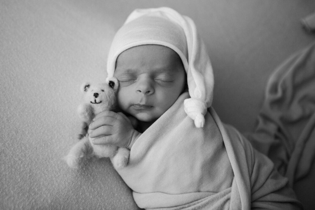 A close up of a newborn baby who is wrapped, holding a little teddy bear with a hat on his head